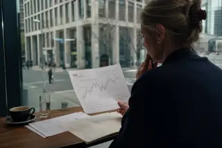 A woman reviews DAX chart printouts at a café window in Frankfurt, city reflections and European financial buildings visible through glass, natural grey daylight.