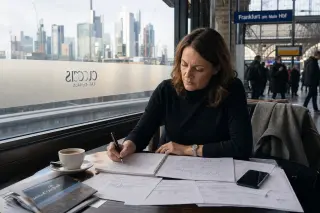 Woman in a European station café reviewing financial documents, sunlight creating soft reflections, subtle urban financial backdrop.