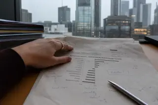 Close-up of a woman's hand on an annotated financial report, with raindrops and Frankfurt city buildings reflected in an office window, conveying thoughtful financial analysis in a minimalist European office.